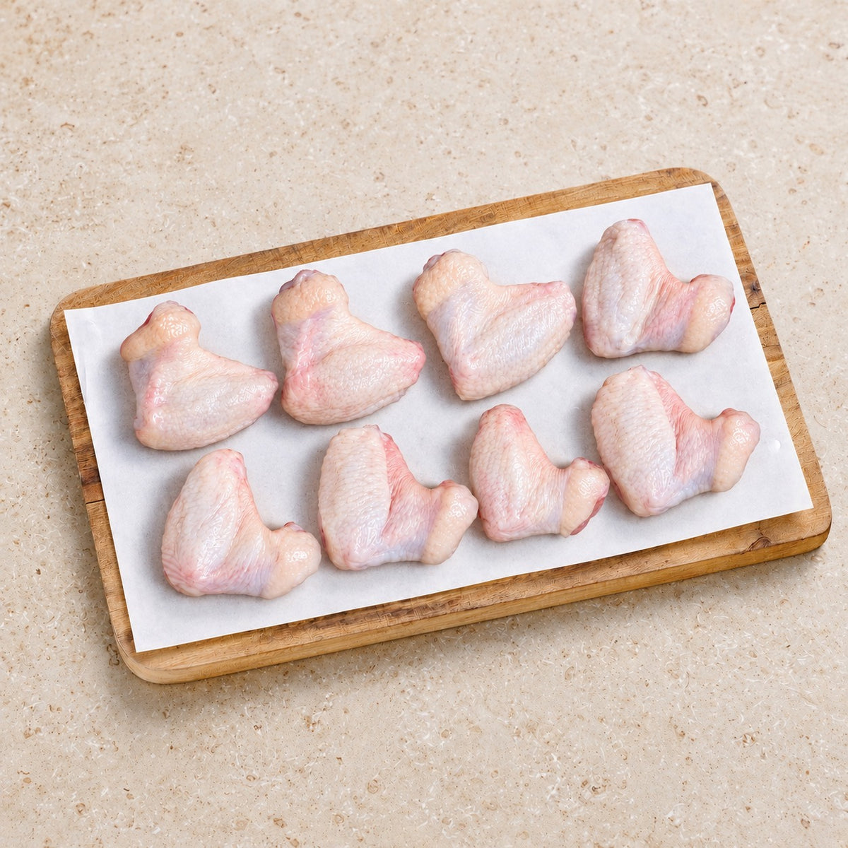Eight raw skin-on chicken wings on white butcher paper and a wooden board on limestone.