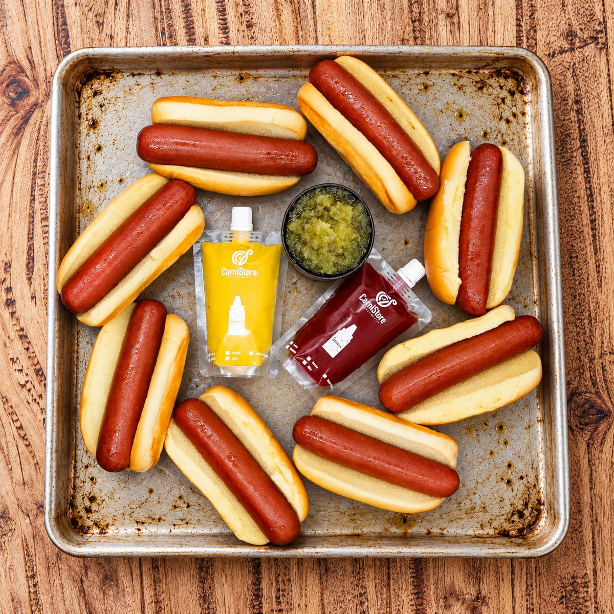 Eight CarniStore smoked Angus hotdogs in potato rolls with mustard, ketchup and relish on a metal tray.