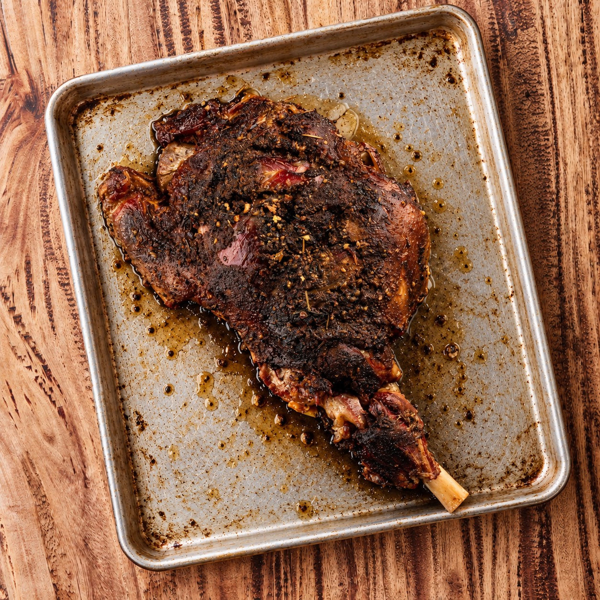CarniStore smoked bone-in lamb leg with a dark spice crust on a silver roasting tray.