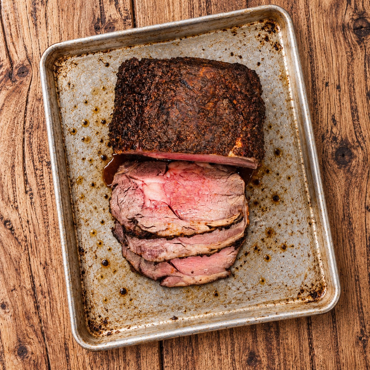 CarniStore smoked ribeye roast with a dark spice crust and pink-centred slices on a silver roasting tray.