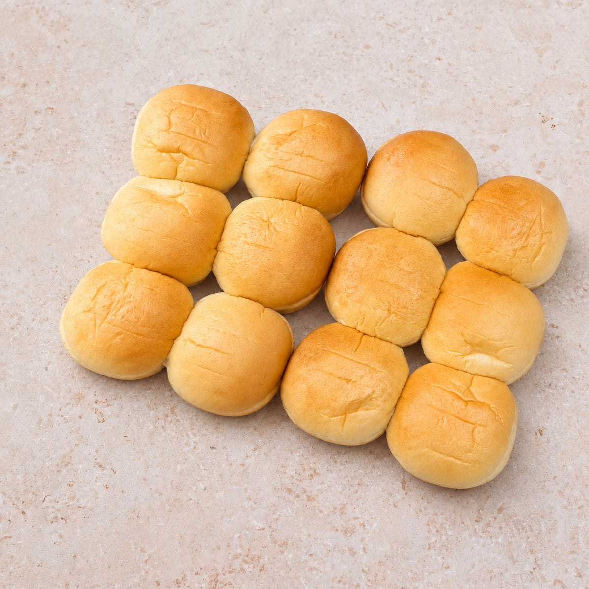 Twelve golden potato slider buns arranged on a light limestone surface.